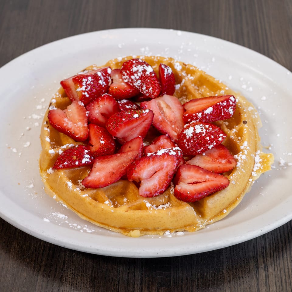 Golden waffle topped with sliced strawberries and powdered sugar on a white plate.