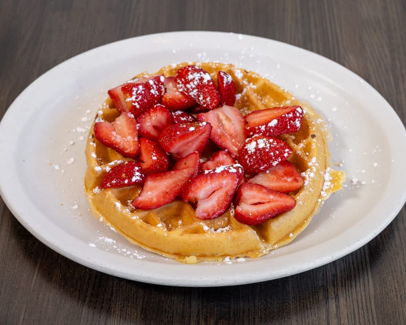 Golden waffle topped with sliced strawberries and powdered sugar on a white plate.