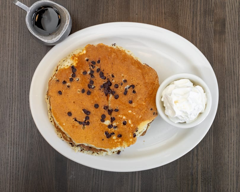 Overhead view of chocolate chip pancakes with whipped cream and a side of syrup.