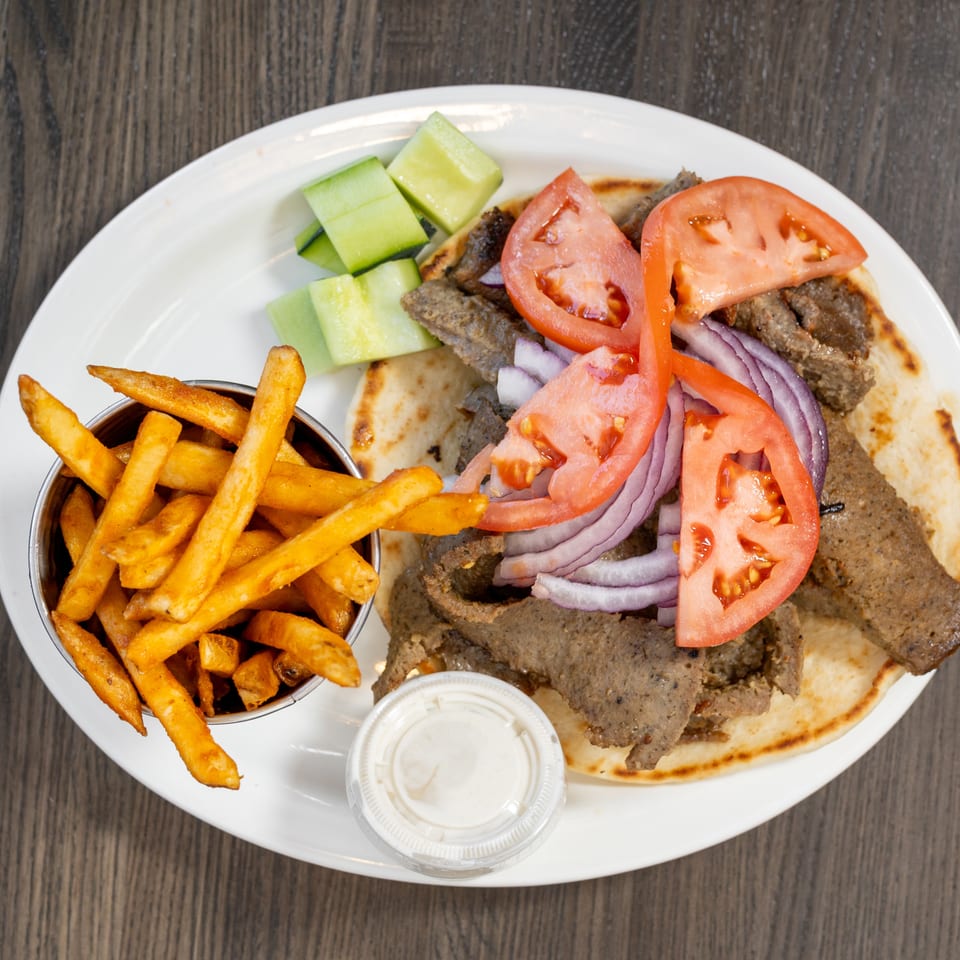 Gyro platter with sliced meat, tomatoes, red onions, cucumbers, fries, and a side of sauce.