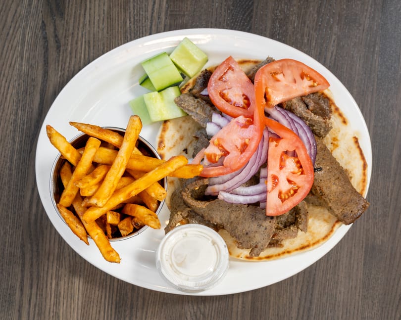 Gyro platter with sliced meat, tomatoes, red onions, cucumbers, fries, and a side of sauce.
