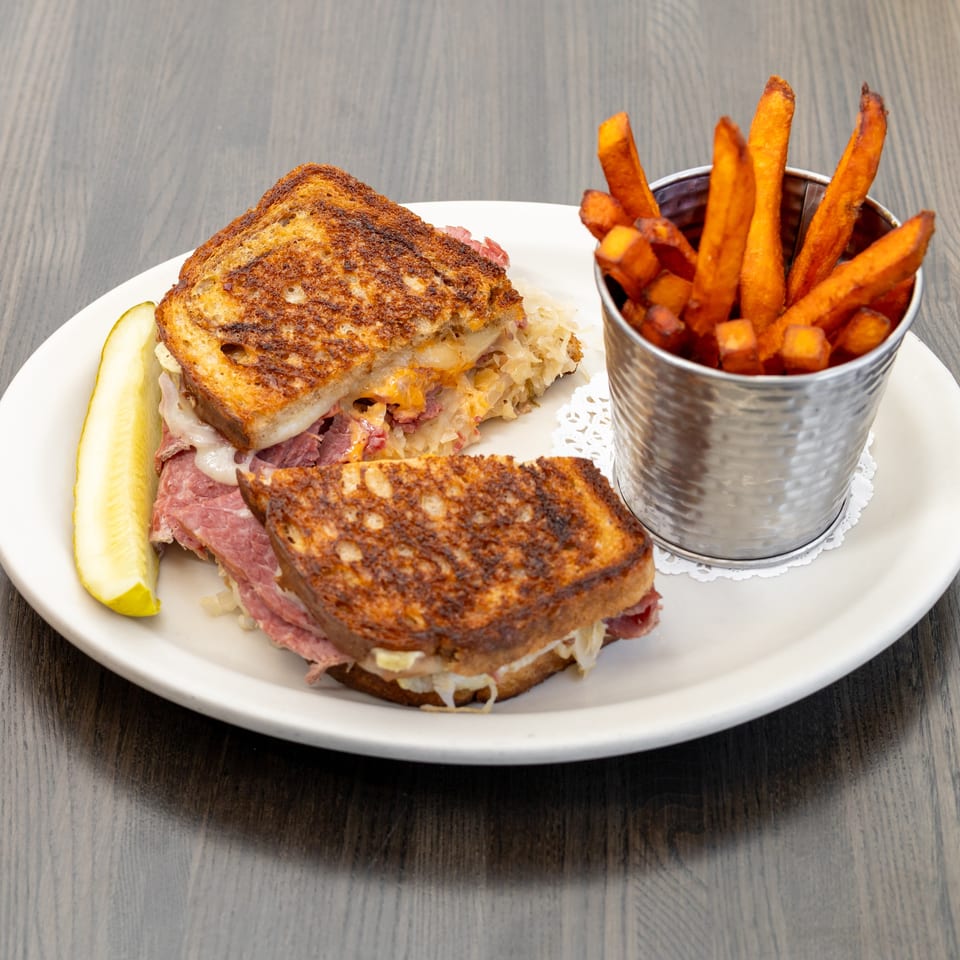Reuben sandwich with corned beef, sauerkraut, cheese, and grilled bread, served with sweet potato fries and a pickle.