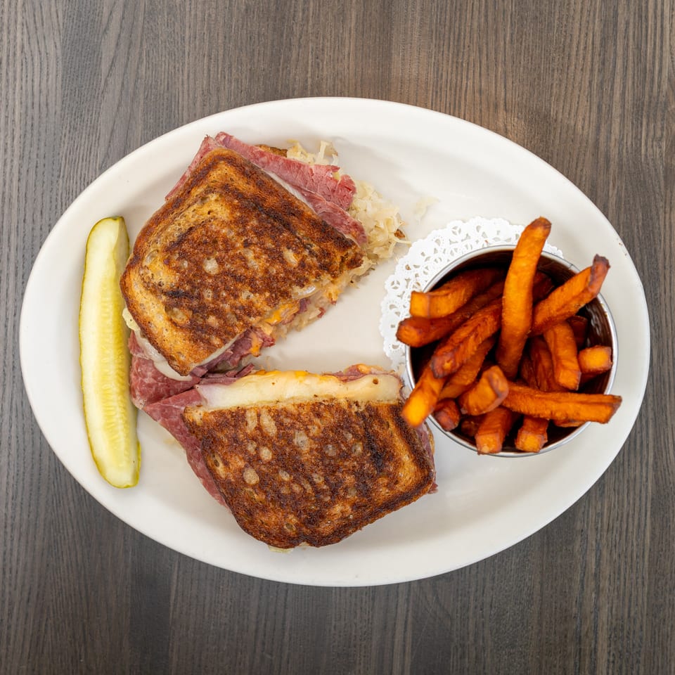 Grilled Reuben sandwich with corned beef, sauerkraut, and cheese, served with sweet potato fries and a pickle.