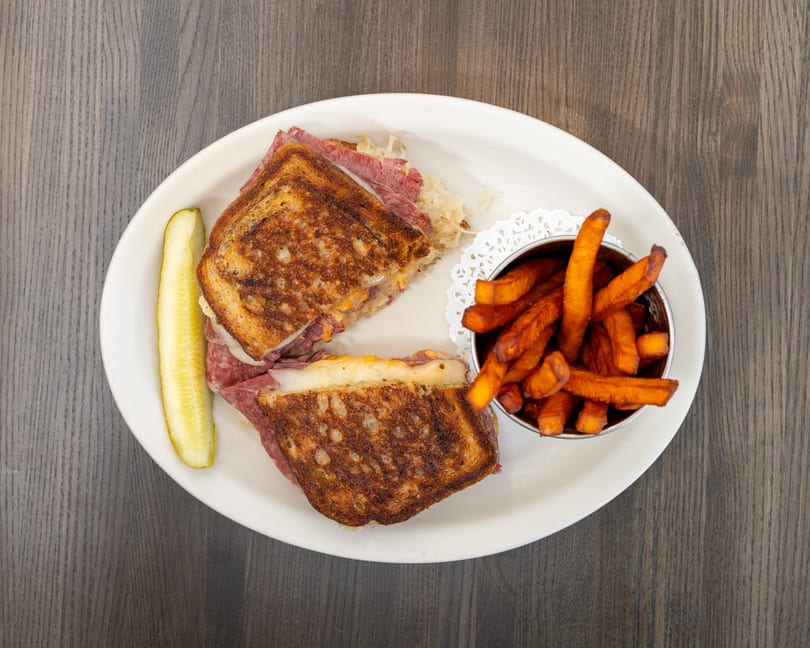 Grilled Reuben sandwich with corned beef, sauerkraut, and cheese, served with sweet potato fries and a pickle.