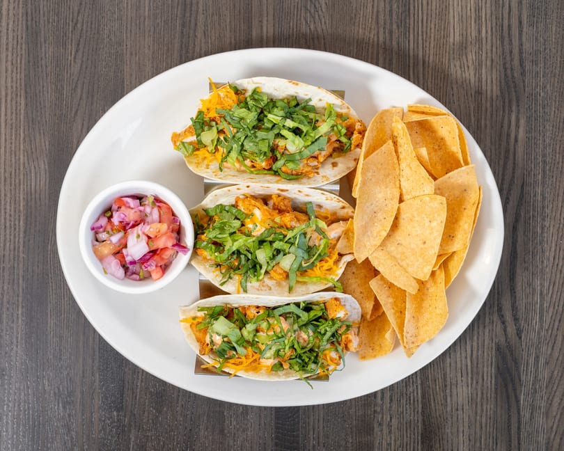 Three chicken tacos with lettuce, cheese, and sauce, served with tortilla chips and salsa.