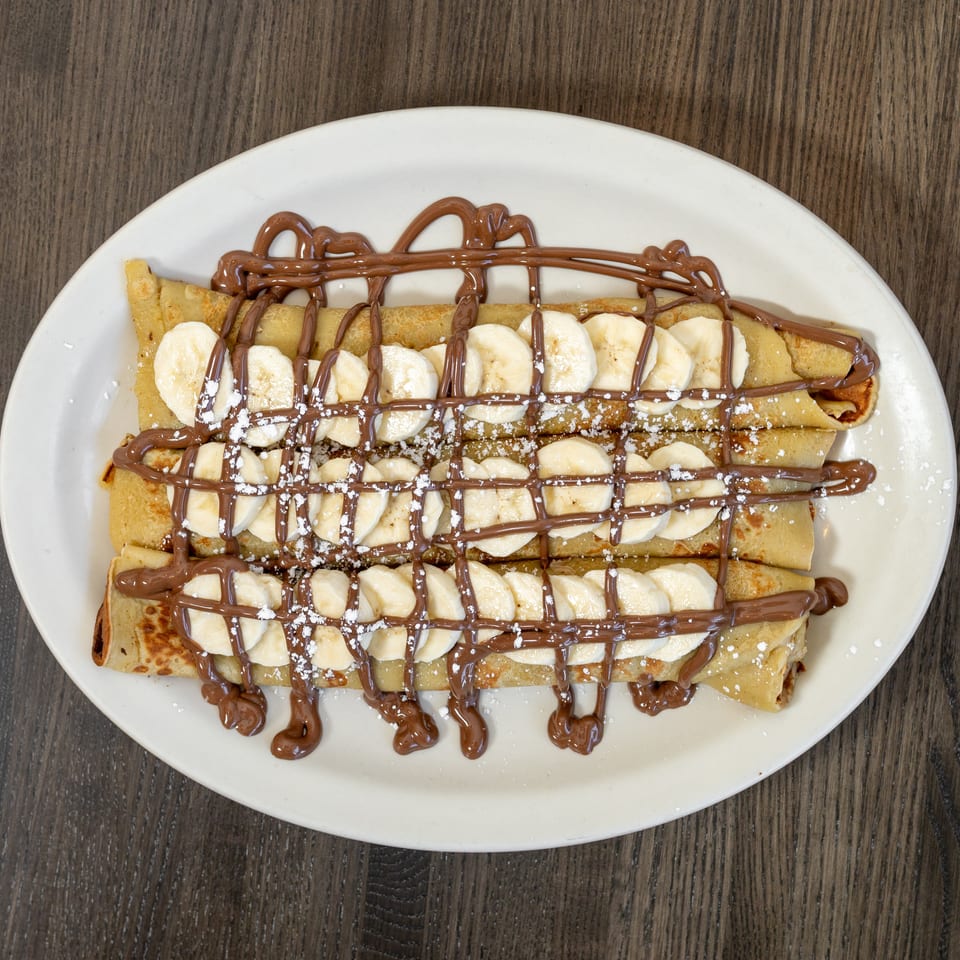Three banana and Nutella crepes, drizzled with chocolate and powdered sugar, on a white oval plate.