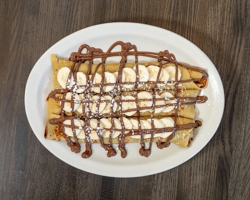 Three banana and Nutella crepes, drizzled with chocolate and powdered sugar, on a white oval plate.