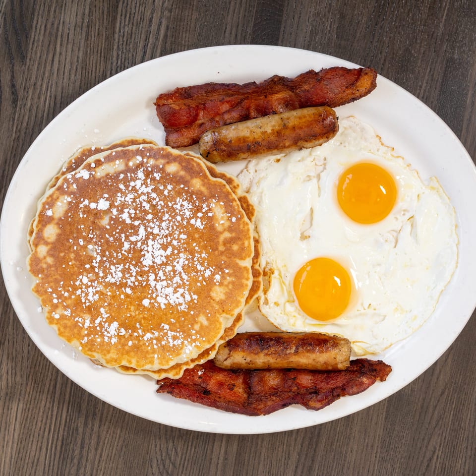 A breakfast platter with powdered sugar pancakes, two fried eggs, sausage links, and bacon strips.