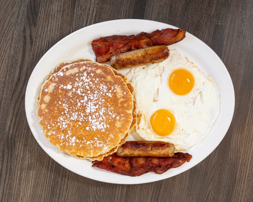 A breakfast platter with powdered sugar pancakes, two fried eggs, sausage links, and bacon strips.